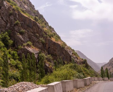 İçinden yol geçen bir dağ sırası. Yol ağaçlar ve kayalarla kaplı. Gökyüzü bulutlu ve güneş parlıyor