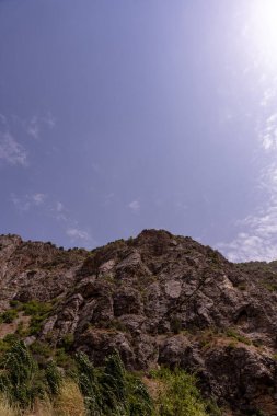 Üzerinde açık mavi gökyüzü olan bir dağ sırası. Gökyüzü bulutlarla kaplı, ama çoğunlukla dağınık ve çok kalın değiller. Dağ sırası yemyeşil bitki örtüsüyle kaplı.