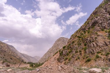 Arka planda bulutlu bir gökyüzü olan bir dağ sırası. Gökyüzü çoğunlukla mavi ve bazı bulutlar etrafa saçılmış. Dağlar ağaçlar ve kayalarla kaplı, manzarayı engebeli kılıyor.