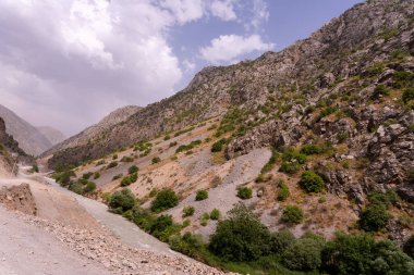 İçinden toprak yol geçen bir dağ sırası. Yol kayalık ve ağaçlar seyrek. Gökyüzü bulutlu ve güneş parlıyor