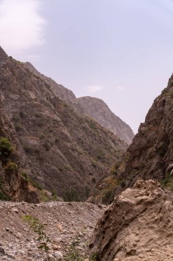 Önünde büyük bir kaya olan kayalık bir dağ vadisi. Gökyüzü bulutlu ve arka planda dağlar var.