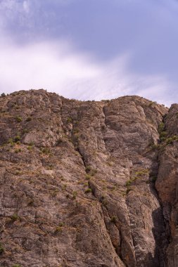 Arkasında bulutlu bir gökyüzü olan kayalık bir dağ. Gökyüzü mavi ve bulutlar beyaz