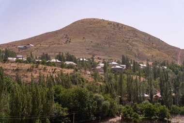 Üzerinde küçük bir köy olan bir yamaç. Tepe ağaçlarla kaplı ve gökyüzü açık.