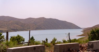 Bir çit bir su kütlesinin önündedir. Su sakin ve berrak. Çit tahtadan yapılmış ve yolun kenarına yerleştirilmiş.