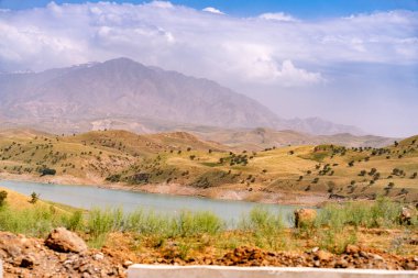 İçinden nehir akan bir dağ sırası. Gökyüzü bulutlu ve güneş parlıyor