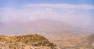 Arka planda bulutlu bir gökyüzü olan bir dağ sırası. Gökyüzü açık mavi bir renktir. Dağlar ağaçlarla kaplı ve toprak toprakla kaplı.
