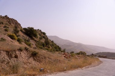 Arka planda yamaç olan bir yol. Tepe kıraç ve kayalık. Gökyüzü açık ve güneş parlıyor.