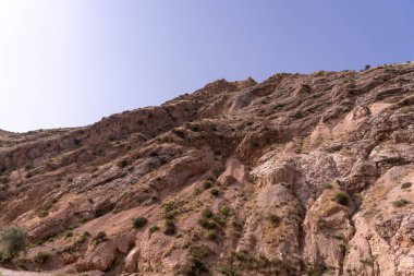 Arka planda açık mavi gökyüzü olan kayalık bir yamaç. Tepe kayalarla kaplı ve seyrek bir bitki örtüsü var. Gökyüzü parlak ve berrak, ortama huzurlu ve huzurlu bir atmosfer veriyor.