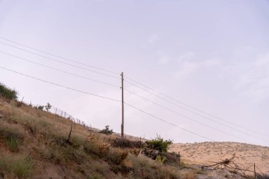 Bir elektrik hattı direği bir alanda duruyor. Gökyüzü bulutlu ve çimenler kuru.
