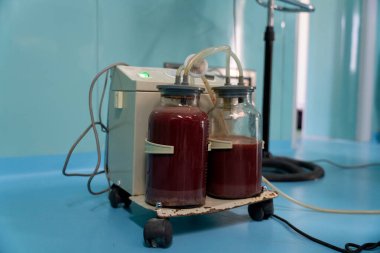 Üzerinde iki şişe kırmızı sıvı bulunan tıbbi bir cihaz. Cihaz tekerleklerin üzerinde ve mavi bir yüzeye yerleştirilmiş.