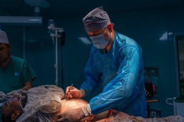 Mavi ameliyat elbisesi giymiş bir adam bir hastanın karnını kesiyor. Sahne bir hastane ameliyathanesinde çekiliyor.