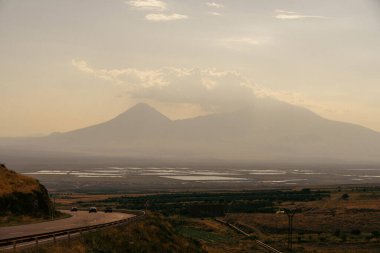 Uzakta bir dağ sırası görülebilir, bir araba yolda ilerliyor. Gökyüzü bulutlu ve atmosfer sakin ve huzurlu.