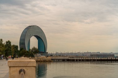 Eğri çatılı büyük bir bina bir şehrin arka planındadır. Gökyüzü bulutlu ve su sakin.