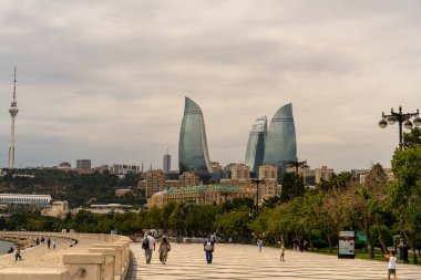 Arka planda uzun bir kule olan bir gökyüzü. İnsanlar su kenarında bir kaldırımda yürüyorlar.
