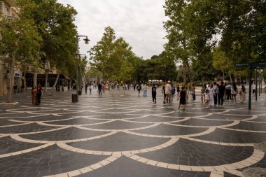 Kalabalık bir cadde, etrafta insanlar dolaşıyor ve arka planda büyük bir ağaç var. Manzara canlı ve hareketli.