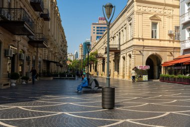 Mavi gömlekli bir adam şehir meydanında bir bankta oturuyor. Meydan boş ve binalar eski.
