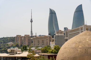 Arka planda uzun bir kule ve tepesinde kubbeler olan iki bina olan bir şehir silueti. Gökyüzü açık ve güneş parlıyor.