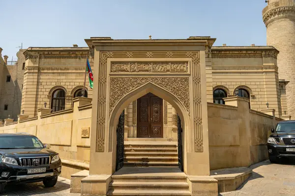 Binanın büyük bir kemer yolu vardır ve taş bir duvarla çevrilidir. Binanın önünde park etmiş iki araba var.