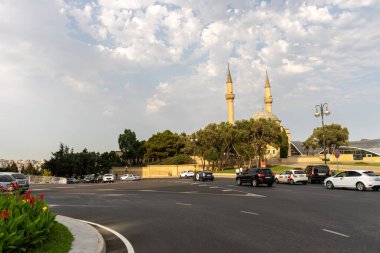Arkasında büyük bir bina olan işlek bir cadde. Gökyüzü bulutlu ve güneş parlıyor