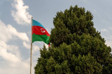 Üzerinde Azerbaycan yazan bir bayrak havada dalgalanıyor. Bayrak mavi, kırmızı ve yeşildir. Gökyüzü bulutlu ve ağaçlar yeşil