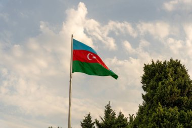 Üzerinde Azerbaycan yazan bir bayrak gökyüzünde dalgalanıyor. Gökyüzü bulutlu ve güneş bulutların arasında parlıyor.