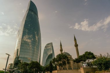 Ortasında yüksek bir bina ve her iki tarafında iki küçük bina olan bir şehir silueti. Gökyüzü bulutlu ve güneş parlıyor