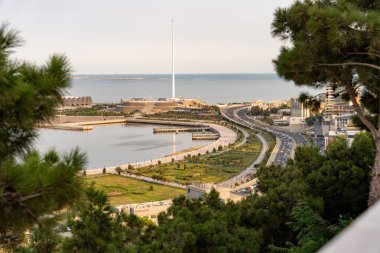 Arka planda büyük bir su kütlesi olan bir şehir manzarası. Şehir hareketlilikle, arabalarla ve sokaklarda yürüyen insanlarla dolup taşıyor. Sahne canlı ve enerji dolu.