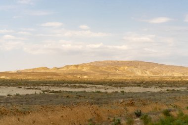 Çorak bir çöl manzarası, birkaç ağaç ve uzak bir tepe. Gökyüzü çoğunlukla açık ve birkaç bulut etrafa saçılmış.