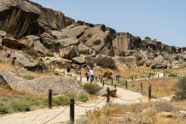 Bir grup insan çölde bir yolda yürüyor. Yol büyük kayalarla çevrili ve arka planda bir çit var.