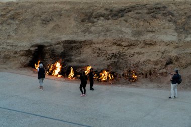Bir grup insan ateşin yanında duruyor, elinde kamera olan bir kişi var. Sahne gergin ve potansiyel olarak tehlikeli, çünkü yangın insanlara yakın ve eğer kontrol edilmezse zarar verebilir.