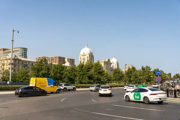 Arka planda büyük bir bina olan işlek bir şehir caddesi. Sokaklar araba ve kamyonla dolu ve etrafta dolaşan bir sürü insan var.