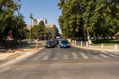 Arka planda kırmızı bir dur işareti olan mavi bir araba caddeden geçiyor. Sokaklar ağaçlarla dolu ve yolda başka arabalar da var.
