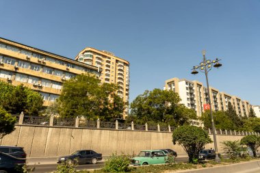Arka planda yüksek bir bina olan bir şehir caddesi. Sokak ağaçlarla dolu ve kaldırım kenarında park edilmiş arabalar var.