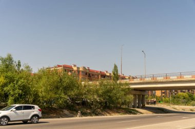 Beyaz bir araba bir köprünün yanındaki caddeden geçiyor. Gökyüzü açık ve güneş parlıyor.