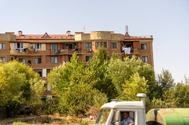 Bir adam büyük bir tuğla binanın önünde bir kamyonun arkasında oturuyor. Binanın bir sürü penceresi var ve etrafı ağaçlarla çevrili.