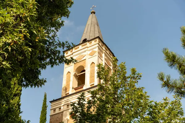 Sivri çatılı ve tepesinde haç olan uzun tuğla bir kule. Kule ağaçlarla çevrili ve arka planda açık mavi bir gökyüzü var.
