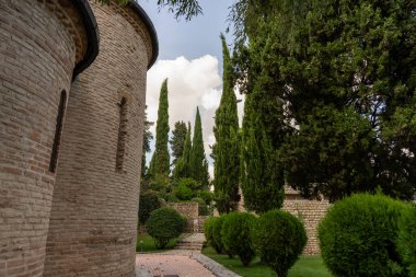 Önünde bir kule ve bir sıra ağaç olan tuğla bir bina. Ağaçlar yeşil ve gökyüzü bulutlu