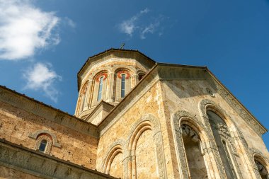 Çan kulesi ve tepesinde haç olan büyük bir bina. Bina eski ve çok fazla detayı var. Gökyüzü mavi ve arka planda bazı bulutlar var