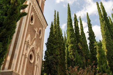Uzun tuğlalı bir kulesi ve etrafını saran çok sayıda ağacı olan bir kilise. Ağaçlar yeşil ve uzun, ve gökyüzü mavi. Sahnenin huzurlu ve huzurlu bir atmosferi var.