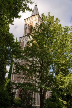 Tepesinde haç ve çan kulesi olan uzun beyaz bir kilise kulesi. Kule ağaçlarla çevrili ve gökyüzü bulutlu.