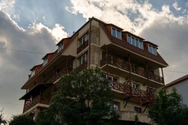 Bir sürü penceresi ve balkonu olan büyük bir bina. Bina ağaçlarla çevrili ve pencerelerden içeri güneş ışığı giriyor.