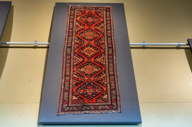 Duvarda mavi arka planı olan bir halı asılı. Halı kırmızı ve mavi ve elmas desenli.