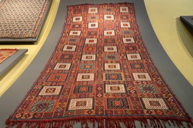 Kare ve üçgenlerden oluşan büyük kırmızı ve beyaz bir halı. Halı diğer halılarla birlikte bir duvarda sergileniyor.