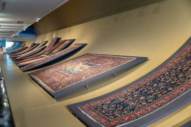 Duvarda bir sıra halı sergileniyor. Halıların boyutları ve renkleri farklı.