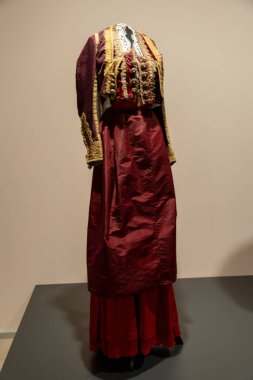 Kırmızı elbiseli ve ceketli bir kadın beyaz bir duvarın önünde duruyor. Kadın kolye ve eşarp takıyor.