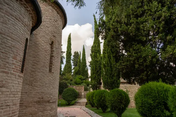 Önünde bir kule ve bir sıra ağaç olan tuğla bir bina. Ağaçlar yeşil ve gökyüzü bulutlu