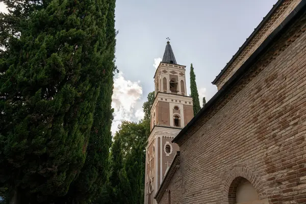 Önünde uzun bir kule ve büyük bir ağaç olan tuğla bir bina. Ağaç yeşil ve bina kahverengi.