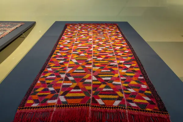 Bir masanın üzerinde geometrik desenli renkli bir halı sergilenir. Halı bir müzede ya da galeride sergileniyor.