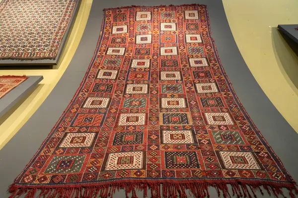 Kare ve üçgenlerden oluşan büyük kırmızı ve beyaz bir halı. Halı diğer halılarla birlikte bir duvarda sergileniyor.