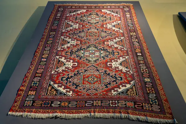 Masada mavi ve kırmızı desenli büyük bir halı oturuyor. Halı çeşitli şekil ve renklerle desenli ve dekoratif bir parça.